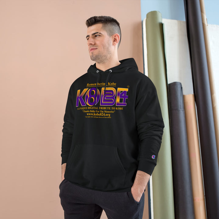 A man wearing a black Champion hoodie with a large purple and gold 'KOBE' graphic on the chest, standing in front of a beige wall with rolled fabric or wallpaper rolls.